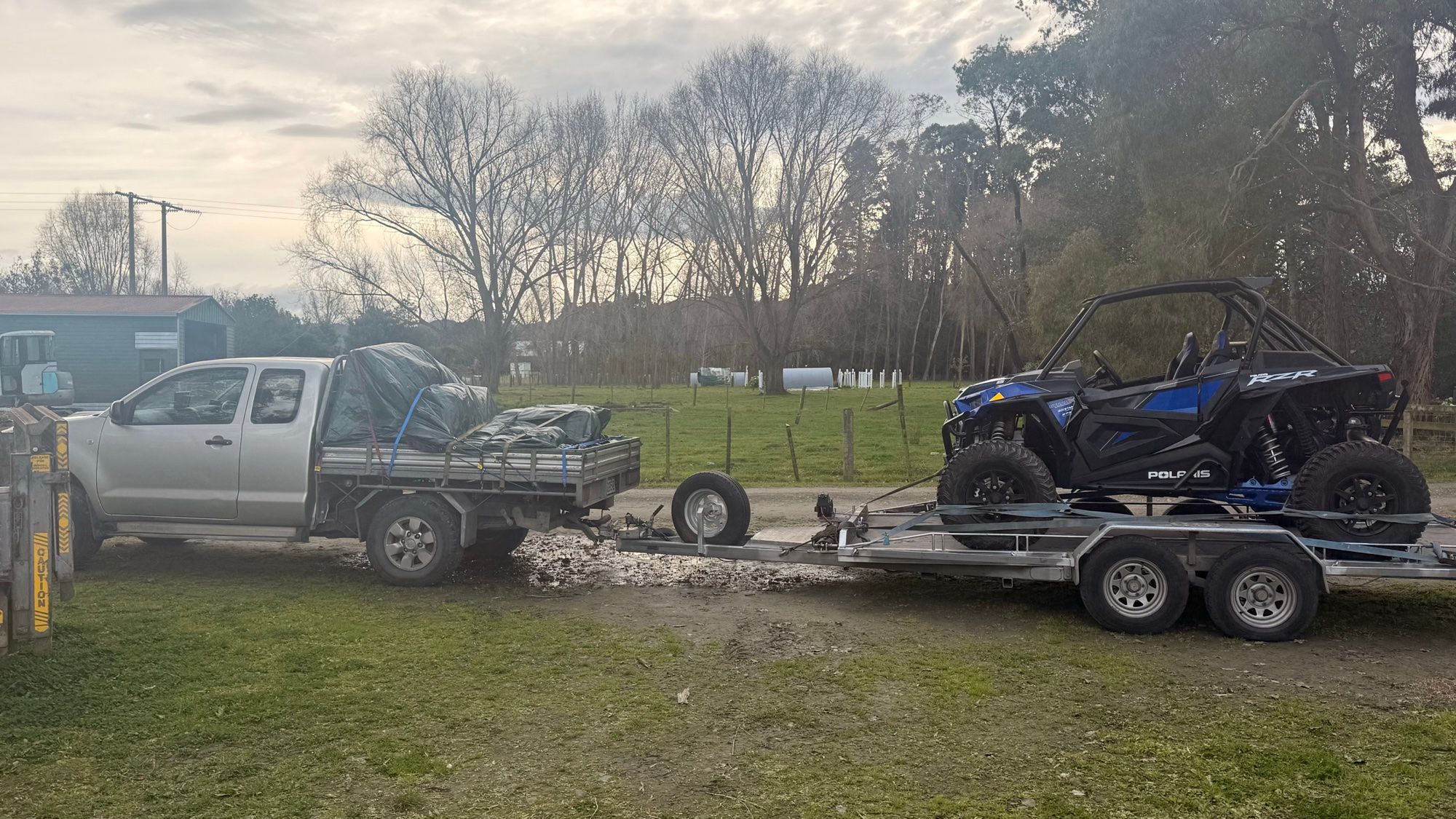 A blue and black Polaris RZR side by side strapped down on the alloy trailer, behind the silver ute with its tray tarped over, on a grass yard.