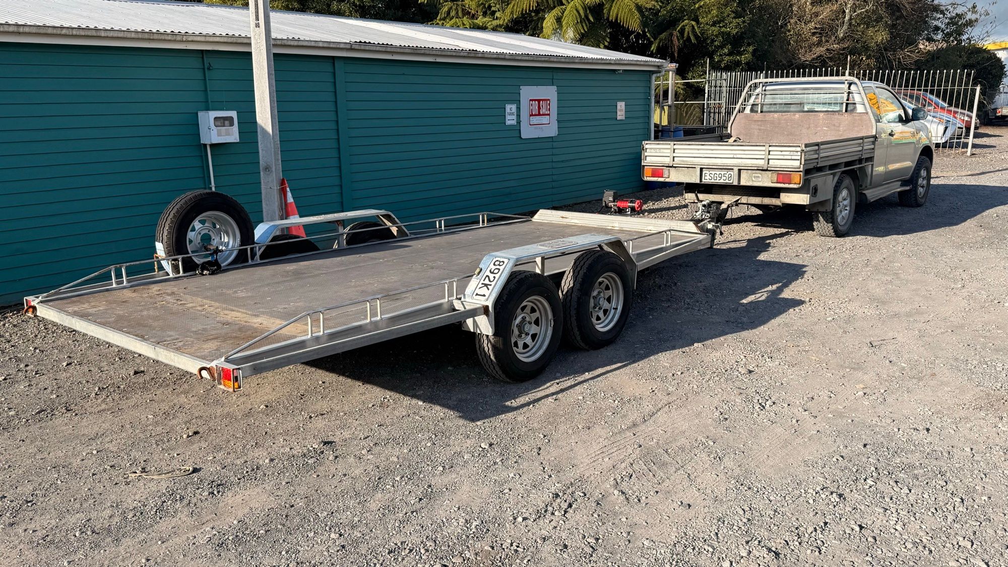 The empty alloy car transporter trailer, tandem axles and side rails, hitched to the silver ute in a gravel yard beside a green shed.